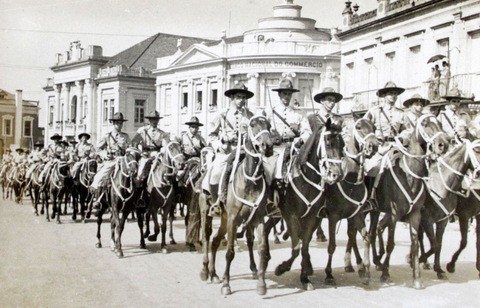 Grupo de homens uniformizados, usando chapéis com abas, montados a cavalo em um desfile na cidade.