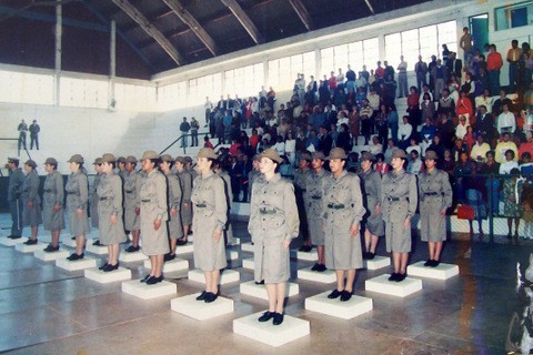 Grupo de soldados, majoritariamente do sexo feminino, uma do lado da outra em um ginásio. Ao fundo um grupo de pessoas em uma arquibancada.