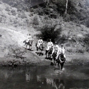 grupo de cinco homens da brigada uniformizados, montados a cavalo, entrando em um rio pela beira.