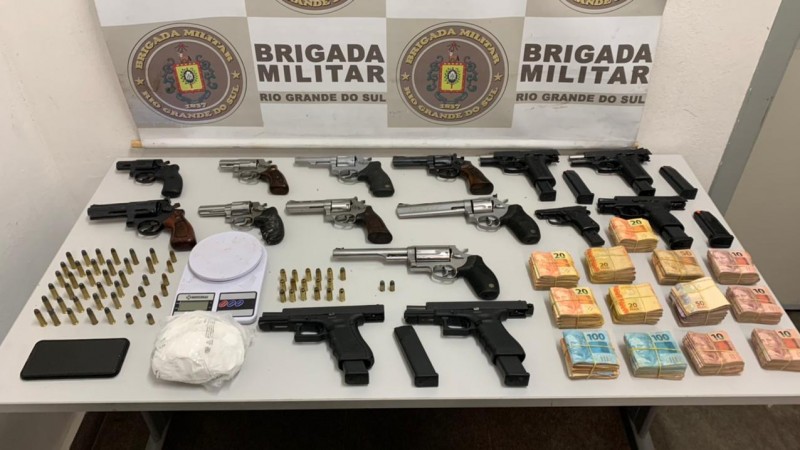 A imagem mostra o material apreendido sobre uma mesa. Ao fundo o banner da Brigada Militar