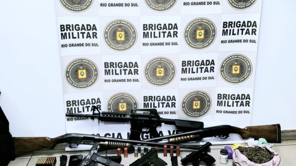 Seis armas, munições, carregadores de celular, porção de maconha e comprimidos estão reunidos em frente a um painel com o Brasão da Brigada Militar. 
