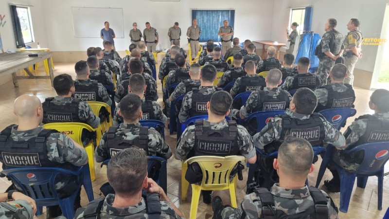 Grupo de homens fardados sentados em cadeiras azuis e amarelas em uma sala branca de costas para a câmera. A frente deles estão militares de pé lado a lado.