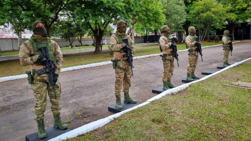 Cinco homens de pé lado a lado, fardados e com o rosto coberto. Nas mãos todos eles portam metralhadoras.