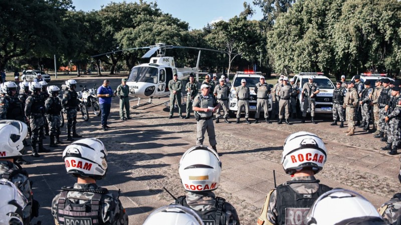 Foto mostra efetivo da Brigada Militar em círculo, com viaturas e helicóptero atrás. No centro, coronel passa orientações ao efetivo.