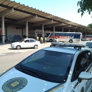 Imagem mostra três policias caminhando até uma estação rodoviária, com duas viaturas paradas próximas ao canteiro central do outro lado, de onde a foto está sendo tirada