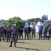 Imagem mostra o coronel Luciano Moritz Bueno, comandante do Policiamento da Capital, cercado por soldados em campo aberto, próximo a um helicóptero.