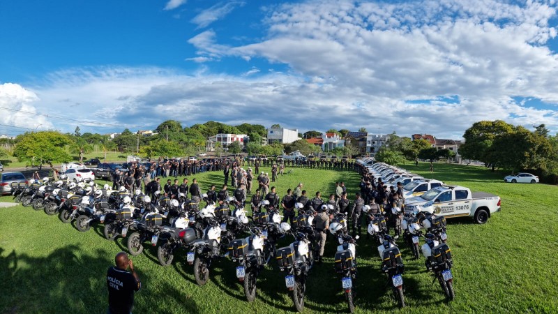 Imagem panorâmica mostra formação de viaturas e policiais militares em um gramado aberto com céu azul