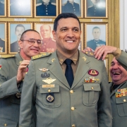 Três militares sorriem enquanto um deles esta recebendo o cargo de comandante