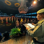 Foto mostra coronel lendo discurso em um ginásio. Os soldados estão perfilados à frente