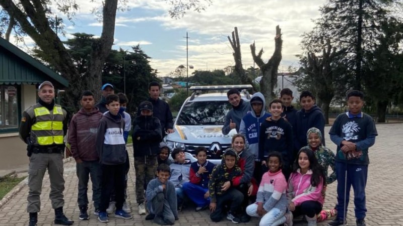 Policial Militar interagindo com crianças e adolescentes do projeto social, todos se encontram próximos de veículo viatura.