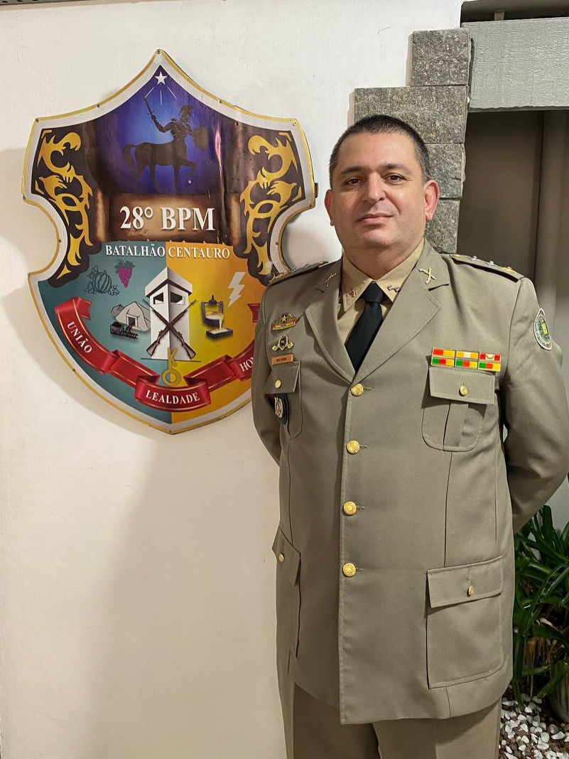 Foto do Comandante do Batalhão ao lado do brasão do 28 BPM