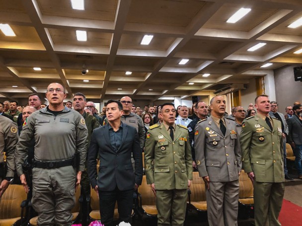 Imagem mostra autoridades perfiladas na primeira fila de um auditório durante execução do Hino Nacional