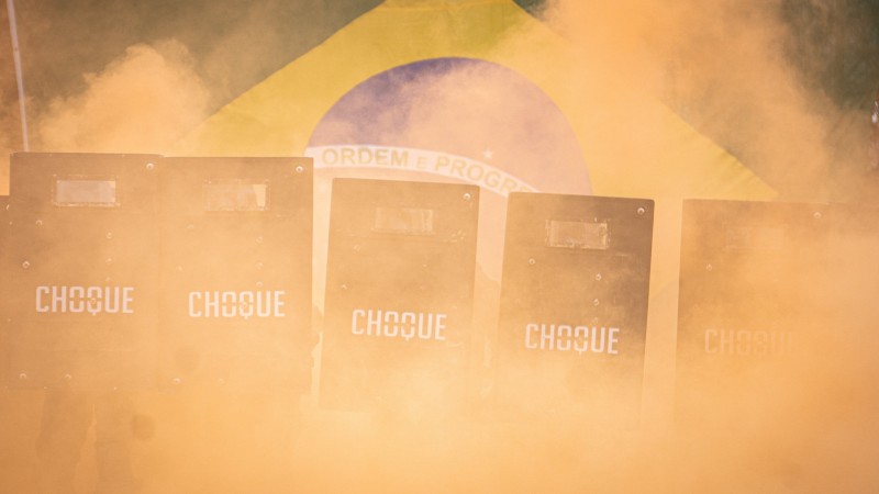 Imagem mostra a entrada dos formandos, que estão segurando escudos do choque em meio à fumaça laranja. Por trás, a bandeira do Brasil