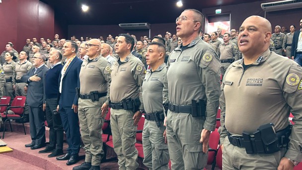 Foto mostra oficiais da BM na primeira fila de um auditório perfilados durante execução do Hino Nacional