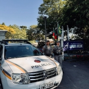 Foto mostra dupla de policiais militares ao lado de uma viatura em frente a uma escola municipal.