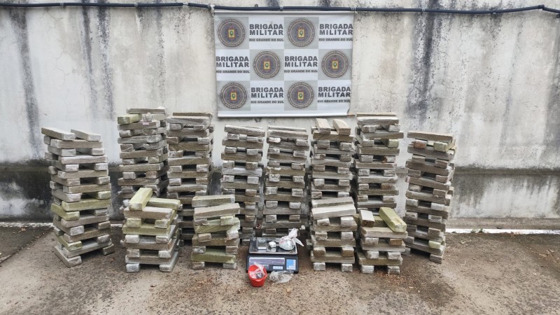 Cerca de 300 quilos de maconha em tijolos empilhados com uma balança e um banner da Brigada Militar ao fundo.