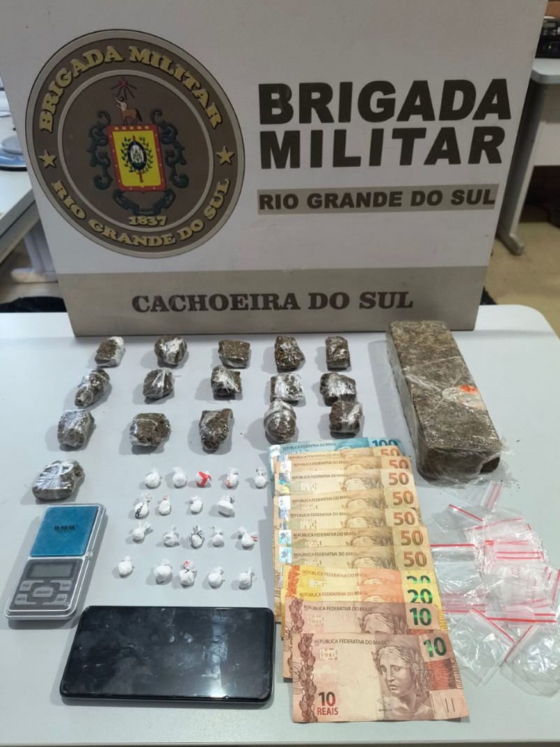 A foto mostra itens apreendidos pela Brigada Militar. Entre os objetos estão embalagens com maconha e cocaína, notas de dinheiro em espécie, uma balança de precisão e um celular, dispostos sobre uma mesa.