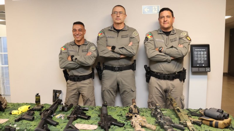 Os tr&ecirc;s comandantes da Brigada Militar aparecem em p&eacute;, com os bra&ccedil;os cruzados, em frente a uma mesa com armas expostas e uma parede clara ao fundo. 
