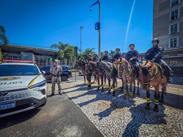 Viatura e PM aparecem ao lado de uma fileira de PMs do Policiamento Montado na Praça Revolução Farroupilha. 
