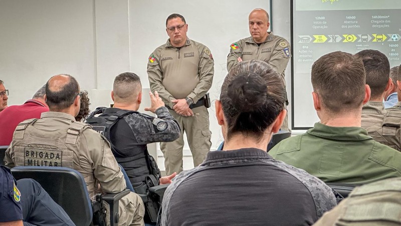 Dupla de PMs aparece em p&eacute; diante de um p&uacute;blico de PMs ouvindo a apresenta&ccedil;&atilde;o do cen&aacute;rio operacional. 