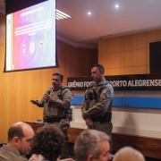 Dupla de comandantes da BM fala diante dos demais representantes de &oacute;rg&atilde;os e institui&ccedil;&otilde;es, com o aux&iacute;lio de um tel&atilde;o, em reuni&atilde;o preparat&oacute;ria.  
