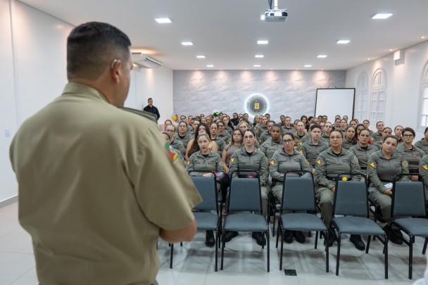As policiais femininas estão sentadas no auditório ouvindo a manifestação do Comandante-Geral da Brigada Militar, que está em pé e de costas. 